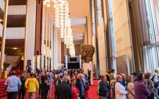 A crowded lobby inside the Kennedy Center, with people walking past a large sculpture and chandeliers under a high ceiling.
